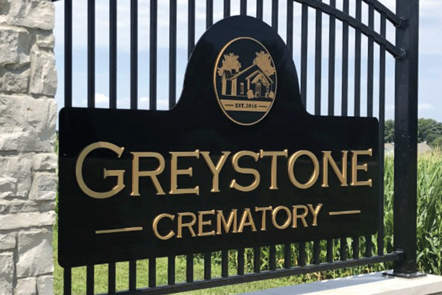 An exterior monument sign for Greystone Crematory installed on a metal gate with a black background, gold acrylic sign letters, and a gold cutout acrylic seal of a building.