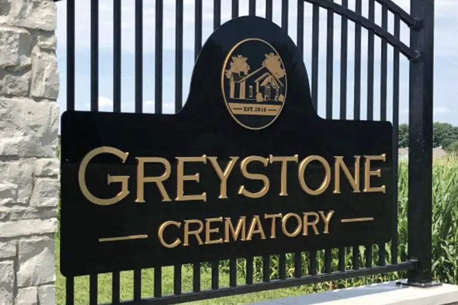 An exterior monument sign for Greystone Crematory installed on a metal gate with a black background, gold acrylic sign letters, and a gold cutout acrylic seal of a building.