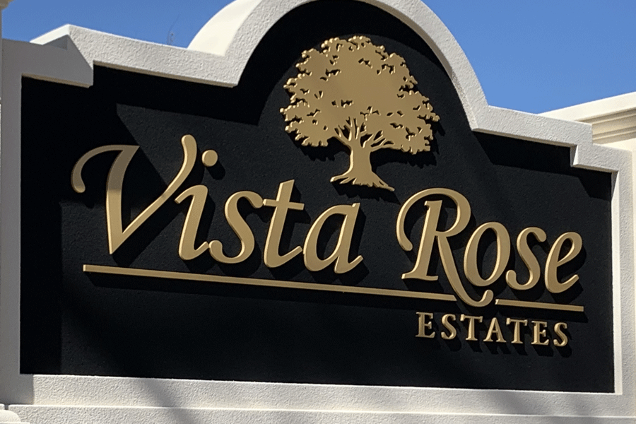 A monument sign for a residential neighborhood called Vista Rose Estates with flat cut gold painted sign letters and a tree on a black background.